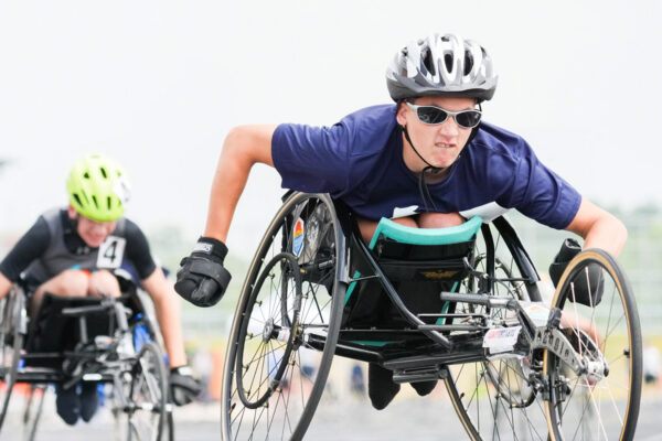Two wheelchair track competitors engaged in a race with the word Inclusion behind them